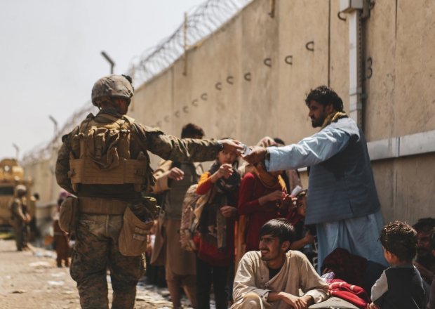 A man in a military uniform hands a bottle of water to an Afghan civilian waiting in a crowd of people.