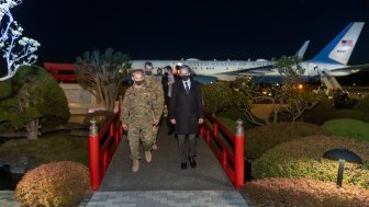 A group of men, some in military uniforms, walk across an ornate bridge at night with an airplane in the background.