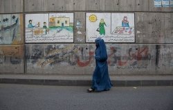 A woman in a blue burqa walks along an empty street in front of murals and graffiti.